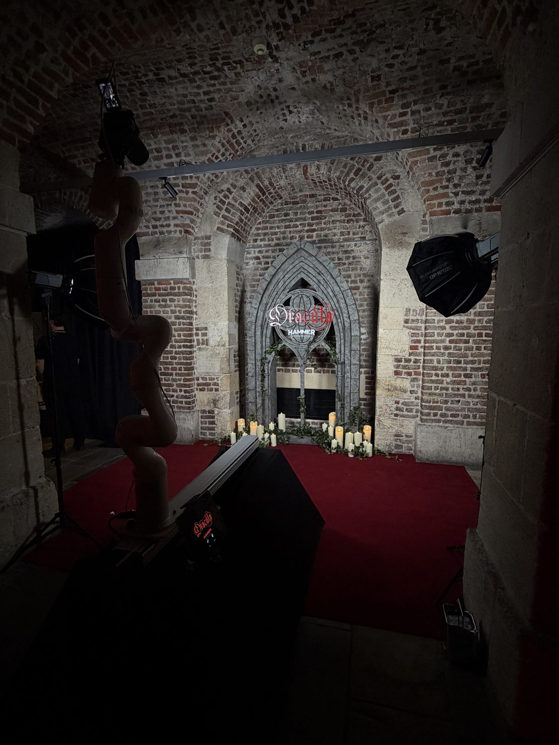 Glambot robot camera ready on the red carpet beside a candlelit gothic Dracula window backdrop in the brick vaults of Café in the Crypt, London